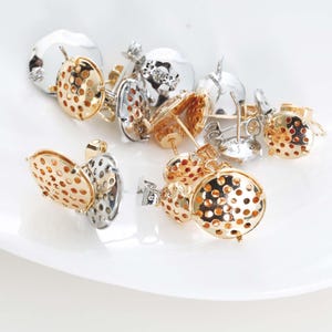 May include: An assortment of gold and silver-toned stud earrings. The earrings feature a round, perforated design, with a post and backing. The earrings are displayed on a white surface, showcasing their metallic shine and intricate details.