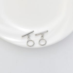 Può includere: Due chiusure a toggle argentate. Ogni chiusura presenta una barra orizzontale collegata a una piccola catena e un anello circolare. Le chiusure sono presentate su una superficie bianca, per la creazione di gioielli.