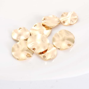 May include: A collection of gold-colored, hammered-look metal charms. Each charm is a rounded shape with a slightly irregular edge and a hole for stringing. The charms are scattered on a white surface.