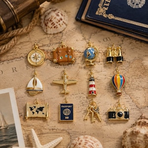May include: A collection of twelve gold-toned metal charms featuring travel-themed designs including a compass, suitcase, globe, binoculars, sailboat, airplane, lighthouse, hot air balloon, map, passport, keys, and a camera with black and white enamel.
