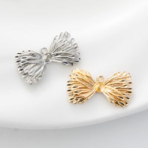 以下が含まれることがあります： 2つの金属製の蝶ネクタイ型のチャーム。1つはシルバー色、もう1つはゴールド色です。各チャームには、ジュエリーに取り付けるためのループが付いています。蝶ネクタイは、テクスチャ加工されたレイヤードデザインです。