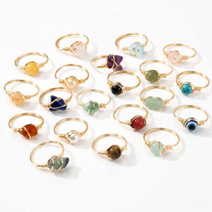 Puede incluir: Una colección de anillos de alambre envuelto con varias piedras preciosas y cuentas en diversos colores, incluyendo azul, verde, rojo, amarillo y blanco. Cada anillo está hecho de un alambre fino dorado que asegura una piedra o cuenta.