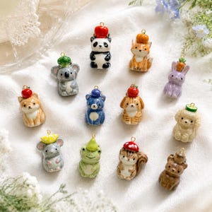May include: A collection of twelve small ceramic animal charms, each featuring a tiny food item on its head. The figures include a panda, fox, koala, rabbit, dog, bear, cat, mouse, frog, squirrel, and other creatures, all equipped with a gold metal jump ring.