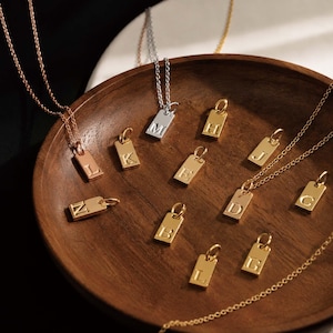 Könnte beinhalten: Eine Sammlung rechteckiger Metallanhänger mit geprägten Buchstaben. Das Set umfasst Gold-, Silber- und Roségoldtöne, wobei einige Anhänger an zarten, passenden Metallketten befestigt sind und in einer runden Holzschale präsentiert werden.