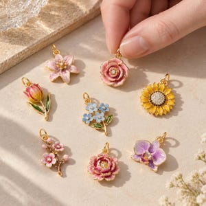 May include: A collection of seven gold-toned metal charms shaped like various flowers, including a pink rose, a yellow sunflower, a purple orchid, a pink tulip, a blue forget-me-not, a pink cherry blossom branch, and a light pink lily, each with a small loop.