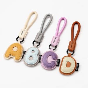 May include: Four letter-shaped keychains featuring the letters A, B, C, and D. Each keychain has a textured faux leather base, a raised letter, a black metal ring, a fabric loop, and a small black tag with the text XXDIAN-life printed in white.