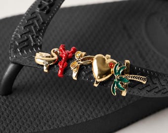 May include: A black flip-flop featuring a textured strap adorned with five metallic charms. The charms include a gold flamingo, a red lobster, a gold bow, a gold heart with a small red stone, and a green and gold palm tree.