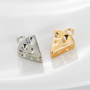 May include: Two cheese-shaped charms, one silver and one gold, each with a loop for attaching to jewellery. The charms have holes to mimic the look of Swiss cheese. The charms are on a white surface.