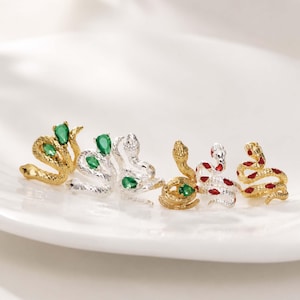 Peut inclure: Un ensemble de six boucles d'oreilles en forme de serpent, déclinées en tons or et argent. Les serpents présentent des corps en métal texturé et sont ornés de petites pierres précieuses en forme de goutte, vertes ou rouges, sur la tête ou le corps.