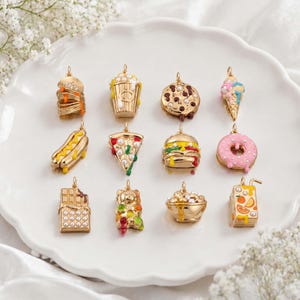May include: A collection of twelve gold-tone metal charms shaped like food items, including a stack of pancakes, popcorn, a cookie, an ice cream cone, a hot dog, pizza, a burger, a donut, a chocolate bar, a gummy bear, cereal, and juice.