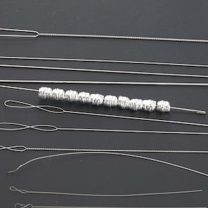 May include: A collection of silver-colored beading needles and wire, some with loops and decorative beads. The needles vary in length and thickness, suitable for jewelry making and crafts.