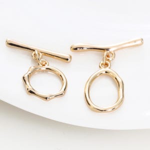 May include: Two gold-colored toggle clasps. Each clasp features a horizontal bar connected to a decorative ring. One ring has an irregular, textured shape, while the other is oval. The clasps are displayed on a white surface.