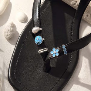 Op de afbeelding: Een zwarte teensandaal met een vierkante neus en een gladde band versierd met vijf decoratieve bedels. De bedels bestaan uit een witte kunstparel, een rond blauw bloemenembleem, een zilverkleurige metalen ring, een lichtblauwe bloem en een blauw staafje.
