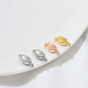 May include: Four jewelry clasps arranged in a row, featuring a smooth, rounded, bean-like shape. The set includes two silver-toned clasps, one rose gold-toned clasp, and one gold-toned clasp, each equipped with small loops at both ends for attachment.