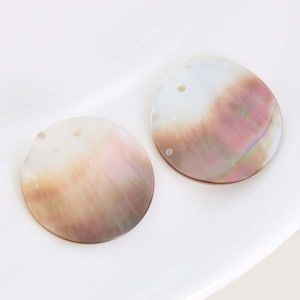 May include: Two iridescent, round shell pendants with a hole drilled at the top for stringing. The pendants have a pearlescent sheen with colors ranging from white to pink and brown. The pendants are on a white surface.