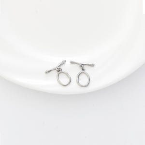 May include: Two silver-toned toggle clasps. Each clasp features a bar and an oval loop. The clasps are displayed on a white surface. These clasps are suitable for jewelry making.