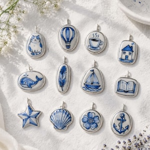 May include: A collection of twelve ceramic pendants featuring blue and white hand-painted designs including a cat, hot air balloon, coffee cup, house, whale, feather, sailboat, open book, star, seashell, flower, and anchor, each with a silver bail.
