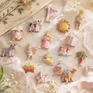 May include: A collection of small ceramic charms featuring various shapes including a pink harp, a spotted cow, a yellow sun, a cherub, a drum, a gift box, a donkey, a cupid, a palm tree, a star, a lantern, a camel, and a white bird.