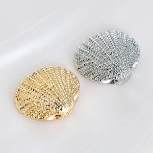 May include: Two seashell-shaped charms. One is gold-colored, the other is silver-colored. Both have a textured surface with a ridged pattern. The charms are likely for jewelry making or other craft projects.