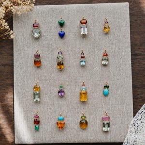 Könnte beinhalten: Eine Sammlung von sechzehn einzelnen Schmuckanhängern, die in einem Raster auf einem hellgrauen Stoff-Display angeordnet sind. Jeder Anhänger besteht aus einer einzigartigen Kombination facettierter Edelsteine in verschiedenen Formen wie Herzen, Ovalen und Rechtecken, gefasst in goldfarbenem Metall mit kleinen Ösen.