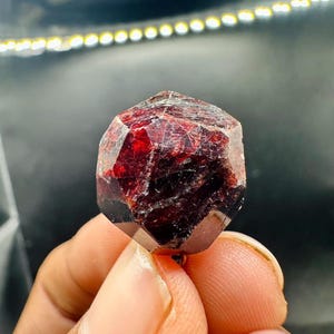 May include: A faceted, deep red garnet crystal with a complex geometric shape. The gemstone is held between fingers, showcasing its rich colour and natural texture. The crystal's surface reflects light, highlighting its internal structure.