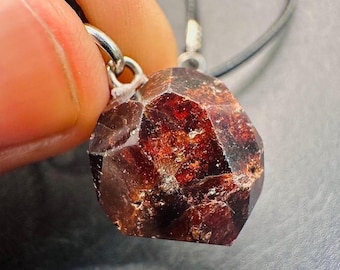 May include: A faceted, reddish-brown gemstone pendant. The pendant is suspended from a gray cord. The gemstone has a rough, natural texture and is held by a silver-colored bail.