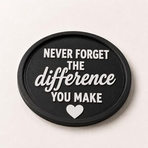 May include: A black, oval-shaped decorative coaster with the text "NEVER FORGET THE difference YOU MAKE" in white lettering. A white heart shape is at the bottom. The coaster has a raised edge.