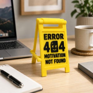 May include: A yellow plastic sign with black text that reads "ERROR 404 MOTIVATION NOT FOUND." The sign has a small graphic of a file with an X through it. The sign is on a wooden desk with a laptop, notebook, and coffee mug in the background.