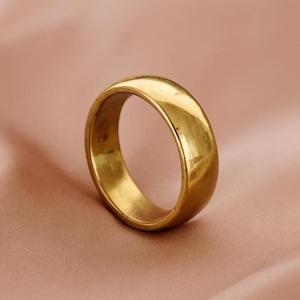 Puede incluir: Un anillo dorado con una banda lisa y redondeada. El anillo está hecho de un material metálico brillante y se encuentra sobre un fondo rosa suave. El diseño del anillo es simple y clásico, con un acabado pulido.