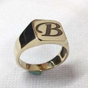 Könnte beinhalten: Ein goldener Siegelring mit rechteckiger Oberfläche. Auf der Oberfläche des Rings ist der Buchstabe "B" in einer Serifenschrift abgebildet. Der Ring ist aus poliertem Gold gefertigt und hat ein klassisches, elegantes Design.