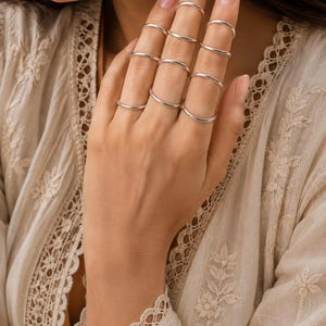May include: A set of nine thin, polished silver-tone metal rings worn across the fingers. Each ring features a simple, smooth, rounded band design with a reflective metallic finish, creating a stacked appearance across the hand.