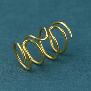 May include: A gold-colored, spiral-shaped ring. The ring has a continuous, flowing design with multiple loops and curves. The metal has a polished, reflective surface, and the ring sits against a teal-colored background.