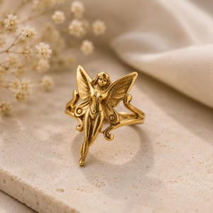 May include: A gold-toned ring featuring a central figure of a winged fairy with detailed wings and a slender form. The metal surface has a polished finish, and the band is integrated into the decorative design of the fairy's outstretched wings.
