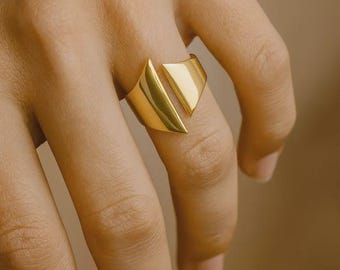 May include: A gold-colored ring with a geometric design. The ring features a split band and a triangular shape on top. The ring is worn on a finger. The ring is made of a shiny metal.