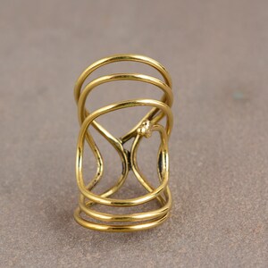 Puede incluir: Un anillo dorado con un diseño único. El anillo presenta múltiples bandas curvas y un intrincado patrón calado. Un pequeño elemento decorativo es visible en la superficie del anillo. El anillo está hecho de un material metálico brillante.