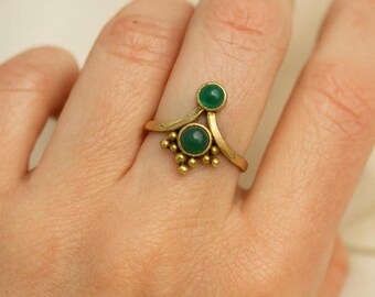 May include: A gold-colored ring with two green gemstones. The ring has a unique design with a curved band and small, round accents. The gemstones are circular and set in the ring's design.