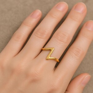 Può includere: Un anello color oro con un design a zigzag. L'anello è indossato su un dito, mostrando la sua forma semplice ma sorprendente. La superficie liscia dell'anello riflette la luce, evidenziando la sua lucentezza metallica. L'anello è un gioiello.