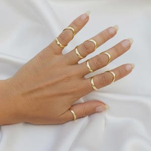 May include: A hand displays several gold rings. The rings include full finger rings and simple bands. The rings are set against a white, silky background, creating a contrast that highlights the jewellery. The rings are likely made of gold.