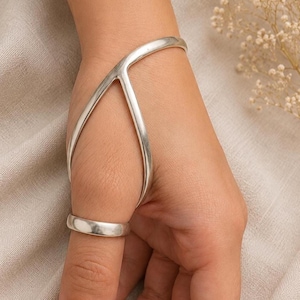 May include: A silver hand bracelet featuring a smooth, polished finish. The piece consists of a curved band that wraps around the wrist and extends across the back of the hand, connecting to a simple, circular ring worn on the thumb.