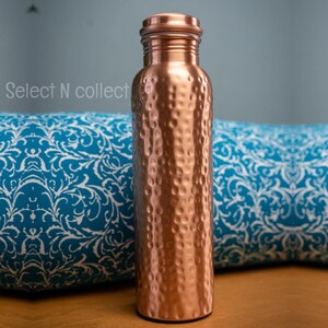 Puede incluir: Una botella de agua de cobre con una textura martillada. La botella tiene una tapa de rosca y una forma cil&iacute;ndrica. La botella se muestra frente a un coj&iacute;n azul estampado. El texto "Select N collect" es visible en el lado izquierdo.