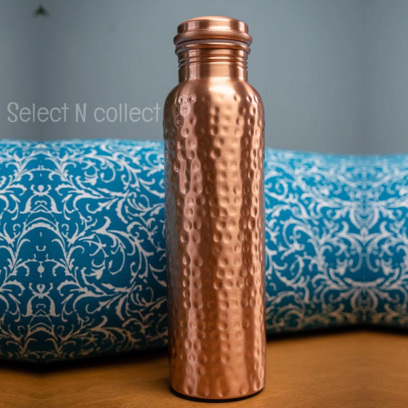 Puede incluir: Una botella de agua de cobre con una textura martillada. La botella tiene una tapa de rosca y una forma cil&iacute;ndrica. La botella se muestra frente a un coj&iacute;n azul estampado. El texto "Select N collect" es visible en el lado izquierdo.