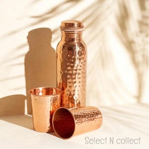 Puede incluir: Una botella de agua de cobre con una superficie texturizada, una tapa a juego y dos vasos de cobre. La botella y los vasos se muestran sobre un fondo neutro con sombras. El texto "Select N collect" es visible.