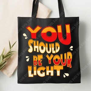 Può includere: Borsa tote nera con la scritta "YOU SHOULD BE YOUR LIGHT" in arancione, rosso e giallo. La borsa ha manici neri ed è esposta su uno sfondo neutro.