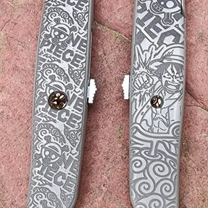 May include: Two silver utility knives with engraved designs. One features a skull and crossbones, the other a character illustration. Both knives have the words "ONE PIECE" etched into the metal, along with decorative swirls.