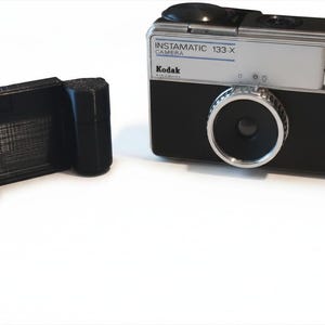 Op de afbeelding: Een vintage Kodak Instamatic 133-X camera met een zilveren en zwarte behuizing, voorzien van een centrale lens en een zoeker, naast een zwart 3D-geprint kunststof accessoire met twee cilindrische zijcomponenten en een plat middenstuk.