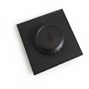 May include: A black square foam pad with a textured surface featuring a circular black plastic lens cap centered on top. The lens cap has a ridged edge and a small embossed logo in the center, providing a secure cover for camera equipment.