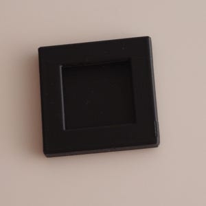 May include: A square black plastic slide mount frame with a hollow center. The frame features a smooth, matte finish and a flat, uniform border surrounding the central opening, designed to hold a photographic slide securely within its structure.