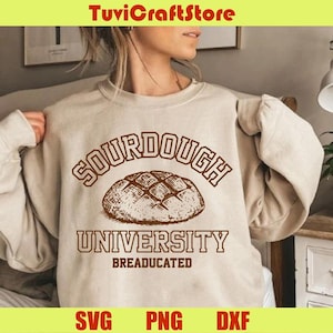 Può includere: Felpa beige con la scritta "SOURDOUGH UNIVERSITY BREADUCATED" in marrone. Un'illustrazione dettagliata di una pagnotta è centrata sopra la parola "UNIVERSITY". La parte superiore dell'immagine presenta la scritta "TuviCraftStore" su uno striscione giallo.