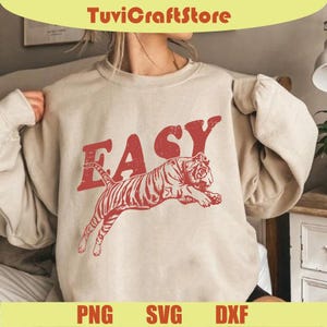 May include: A beige sweatshirt with a red graphic of a tiger leaping and the word "EASY" above it. The sweatshirt has long sleeves and a crew neck. The bottom of the image shows the words "PNG", "SVG", and "DXF".