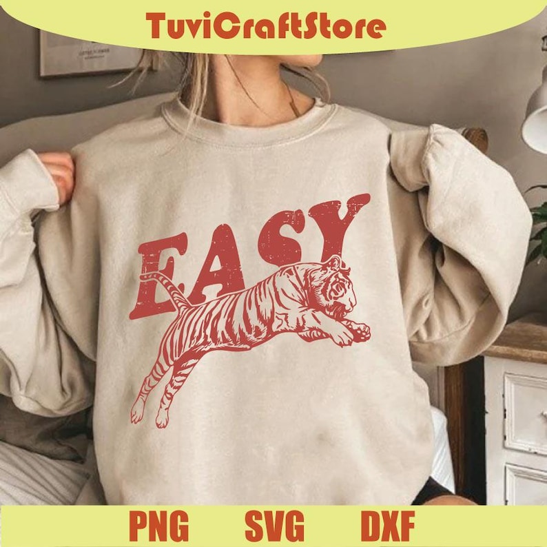 May include: A beige sweatshirt with a red graphic of a tiger leaping and the word "EASY" above it. The sweatshirt has long sleeves and a crew neck. The bottom of the image shows the words "PNG", "SVG", and "DXF".